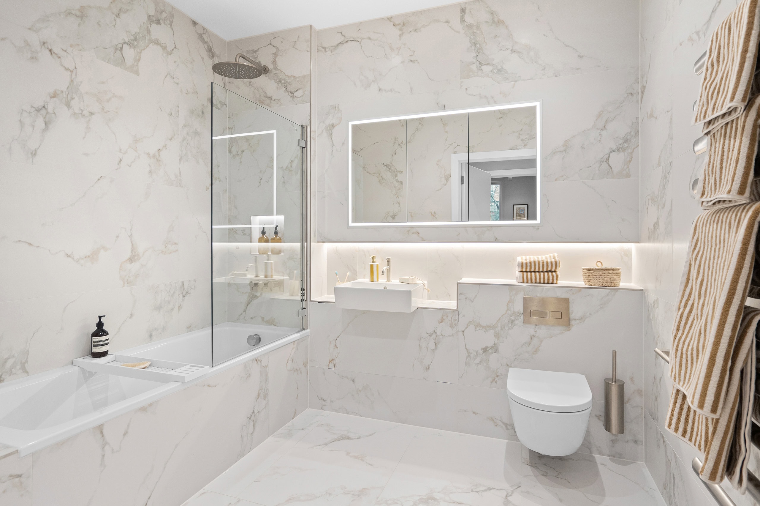 Contemporary bathroom with marble effect porcelain tiles from floor to ceiling. Bath and shower area with fitted screen to the left, to the right is a towel rack with illuminated mirror cabinet on the far wall, with a toilet in the right-hand corner