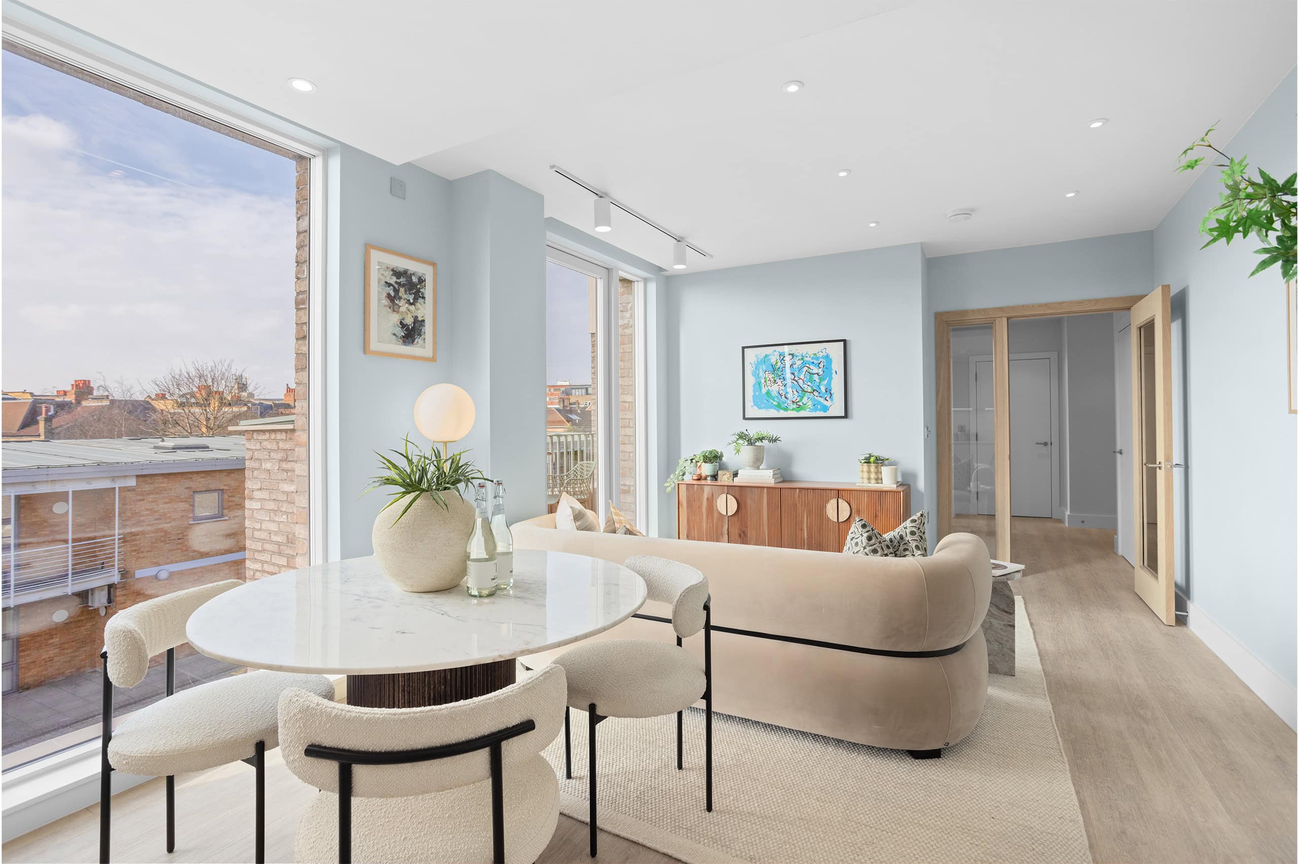 Large, naturally well-lit living area with amtico wood floorboards and floor to ceiling window looking outside to a balcony view. A sofa and kitchen table sit on a large carpet.