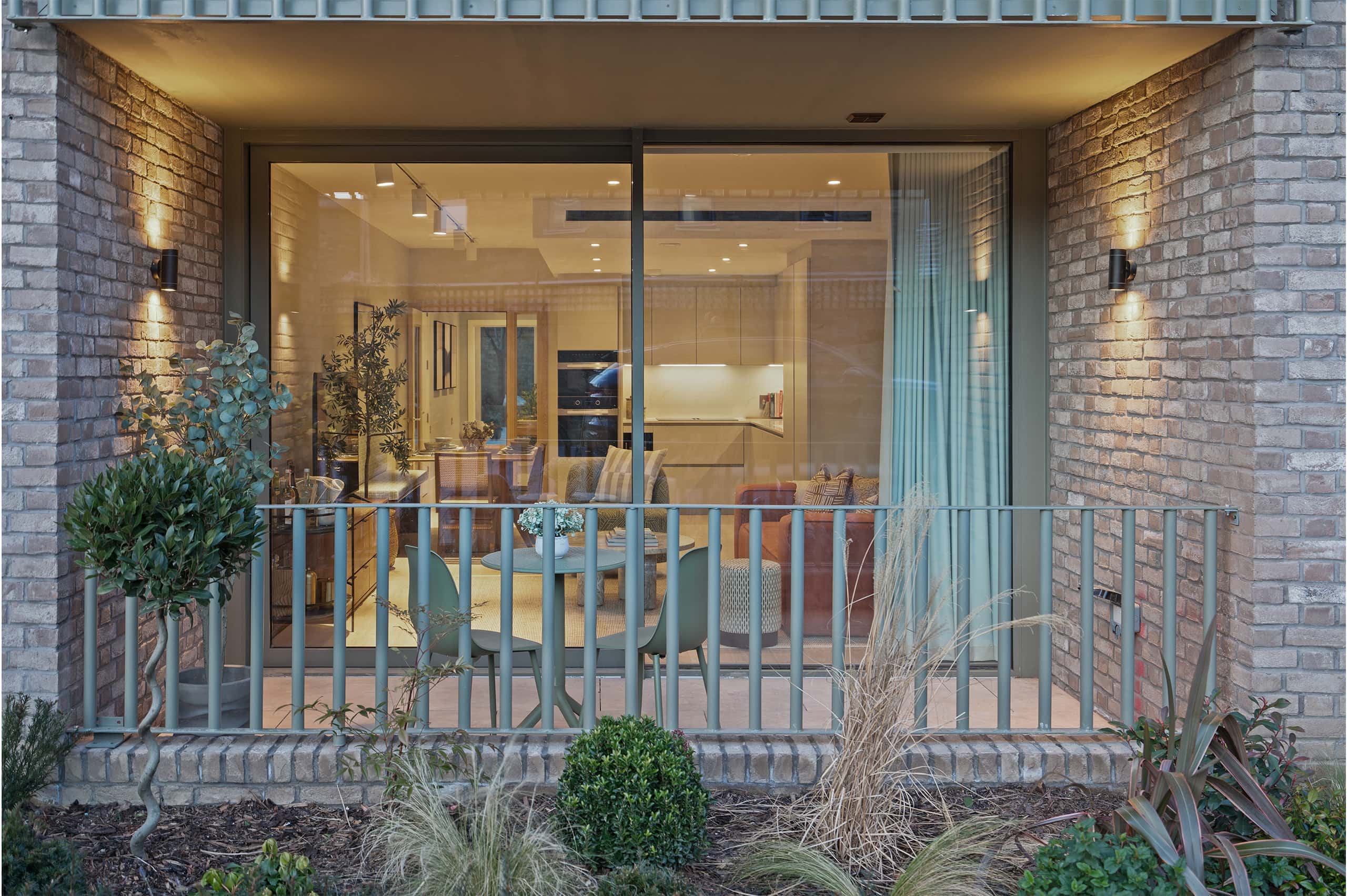 View from the exterior into a warmly lit apartment at night, showcasing a stylish open-plan living space with a modern kitchen and cosy seating area. The private front terrace in the foreground features a small outdoor dining set, soft landscaping, and lush greenery, framed by a sleek metal railing. The apartment’s floor-to-ceiling windows highlight the inviting interior, blending contemporary design with natural textures.