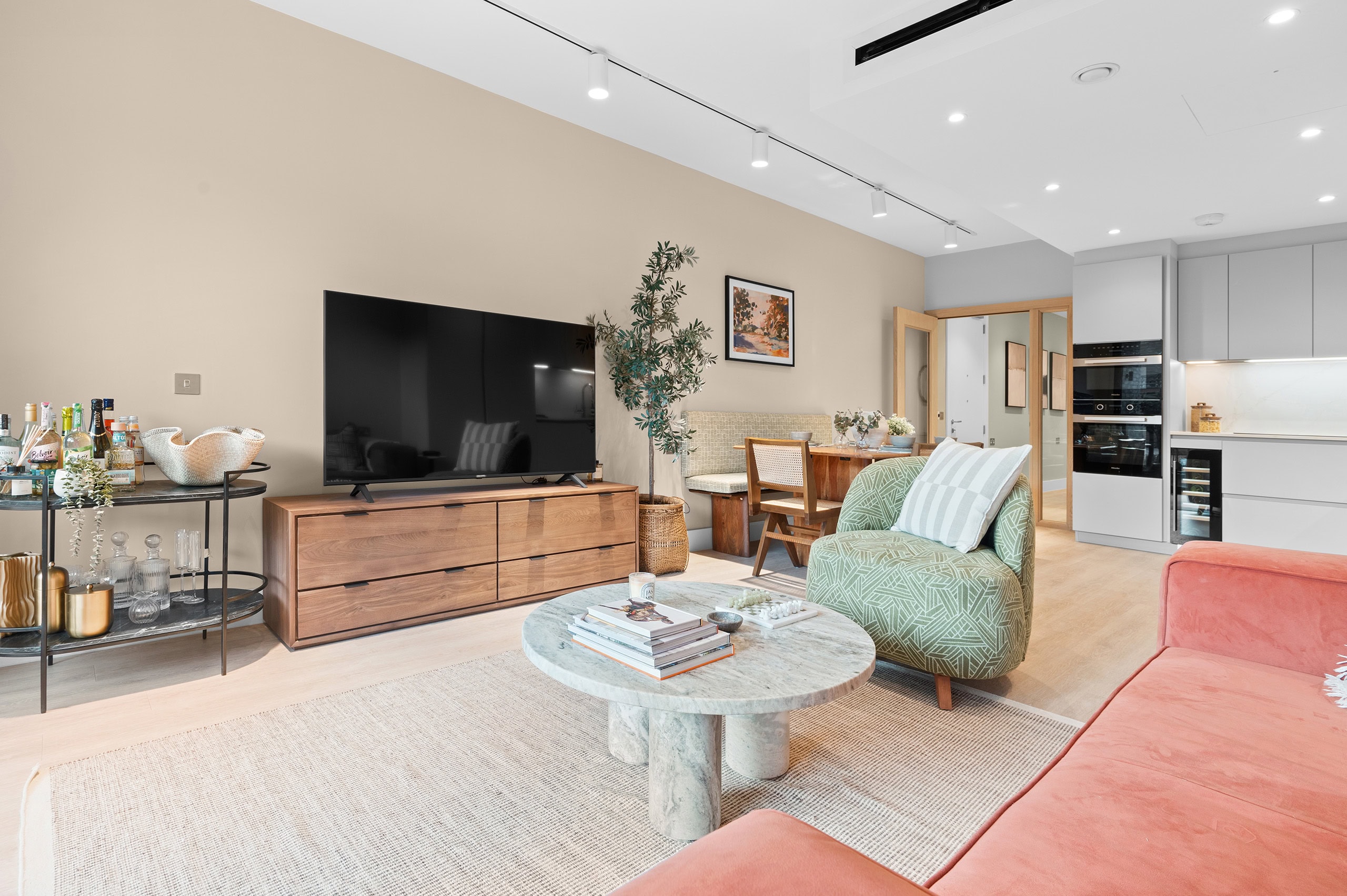 Living/kitchen area showing a large television mounted on a wooden cabinet, a coffee table and fitted kitchen appliances and cupboards in the background